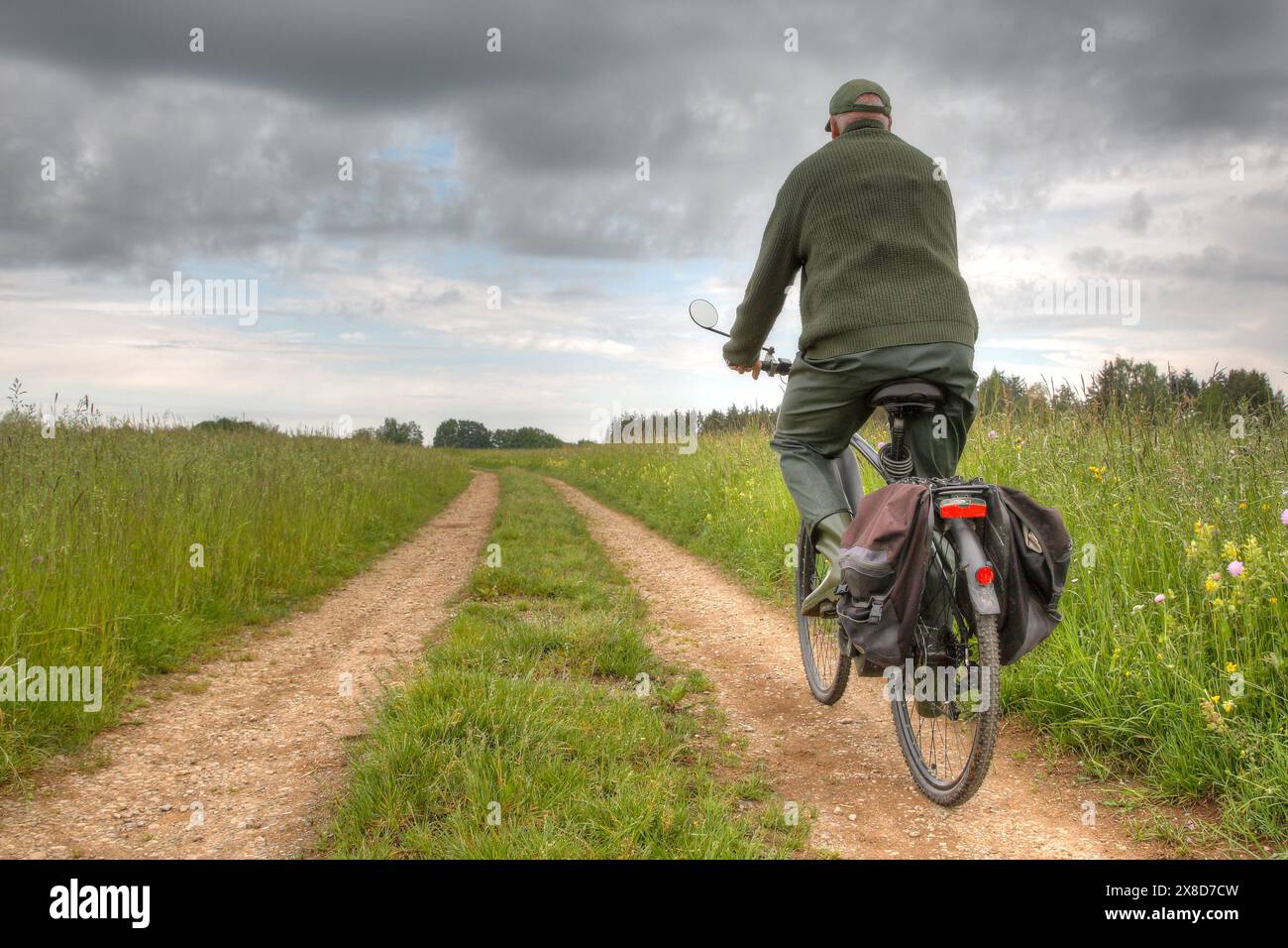 An active senior rides his bicycle on a dirt path between two green meadows. The sky is cloudy and changeable, creating an uncertain atmosphere. Stock Photo