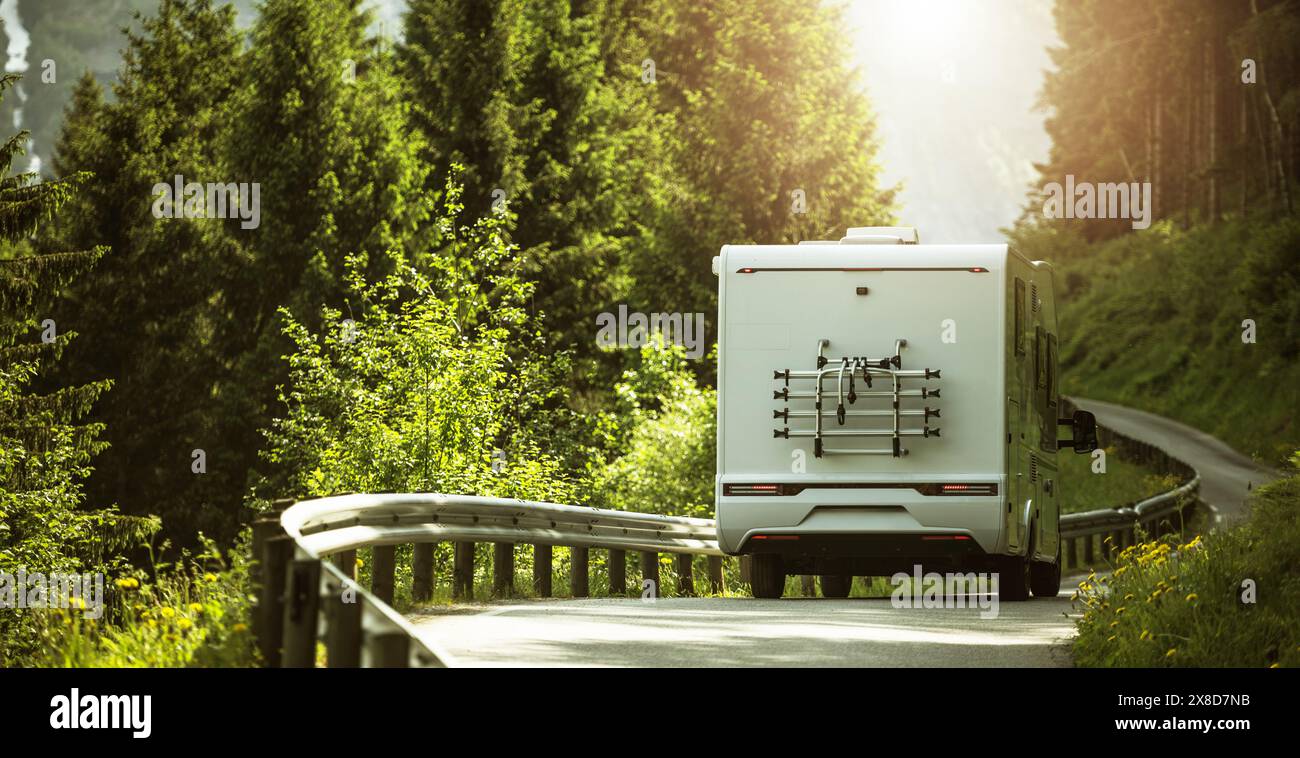 RV camper van is seen driving down a road that runs parallel to a dense forest. The scenario captures the vehicles movement against the backdrop of th Stock Photo