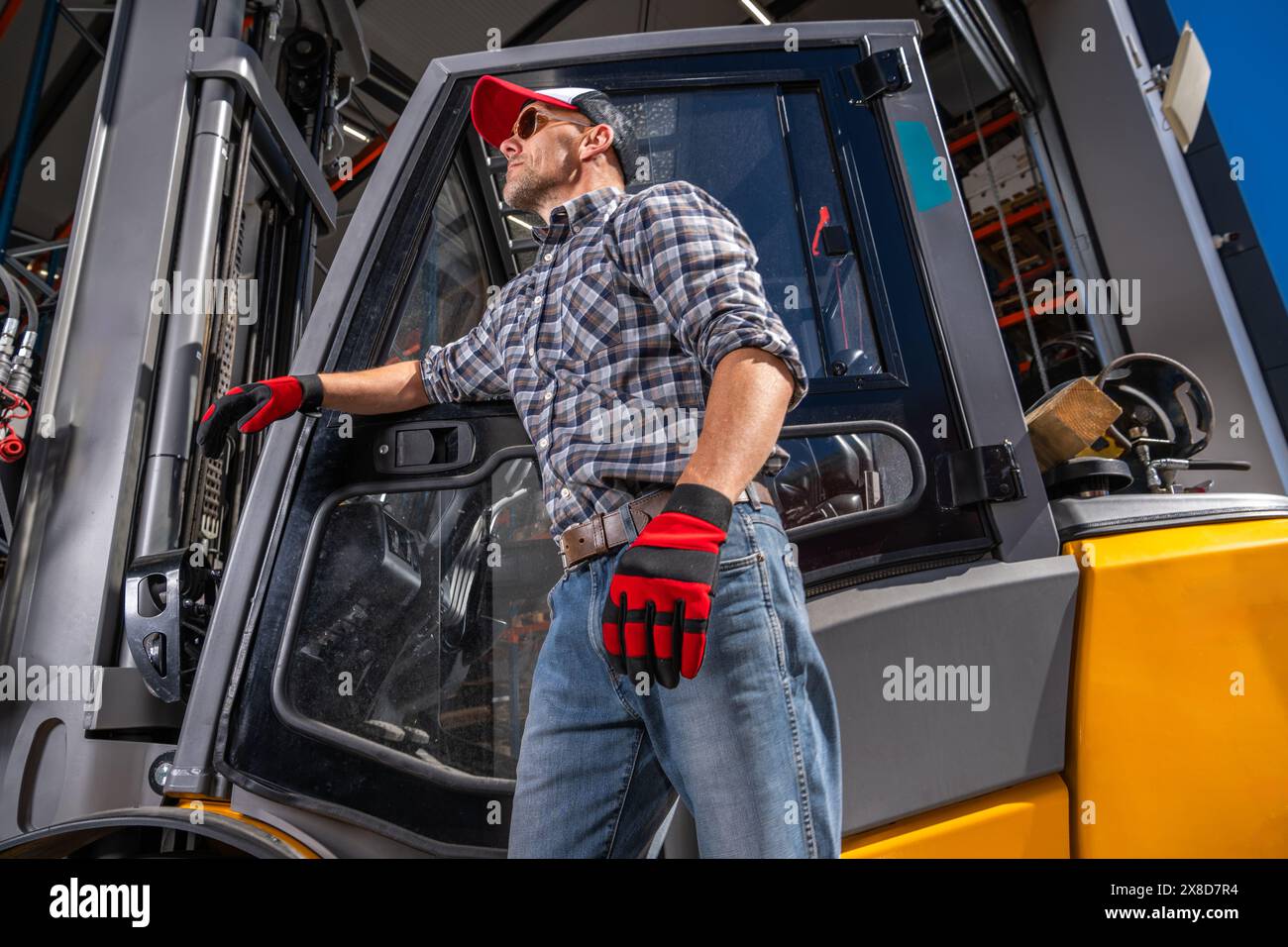 A worker in work attire stands confidently in front of a forklift in an industrial setting. Stock Photo