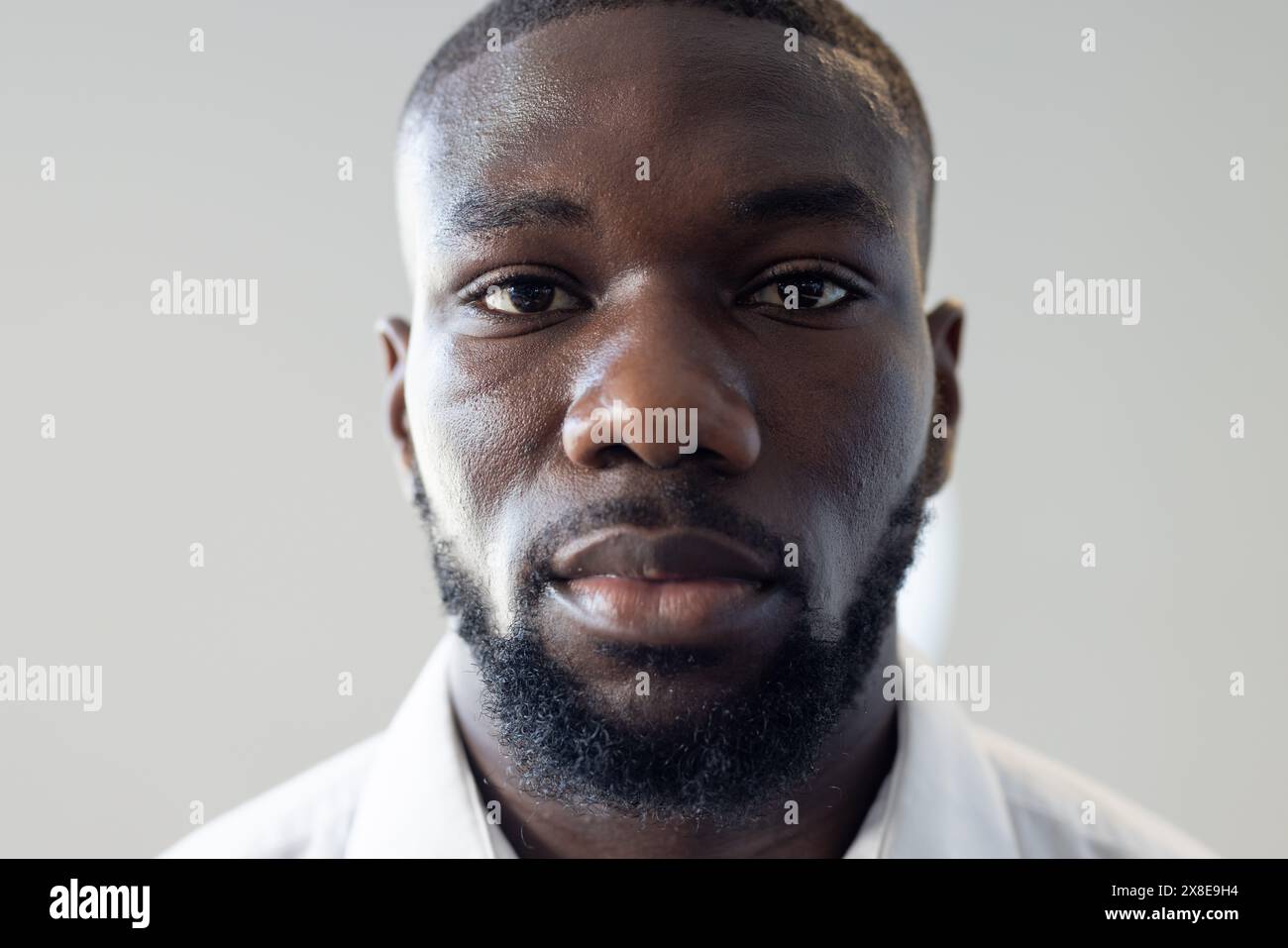 At modern office, young African American man with short hair and beard looks forward. Wearing white shirt and having serious expression on his face, s Stock Photo