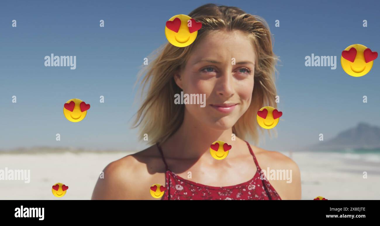 Image of emojis over smiling beautiful caucasian woman walking at beach against sky Stock Photo