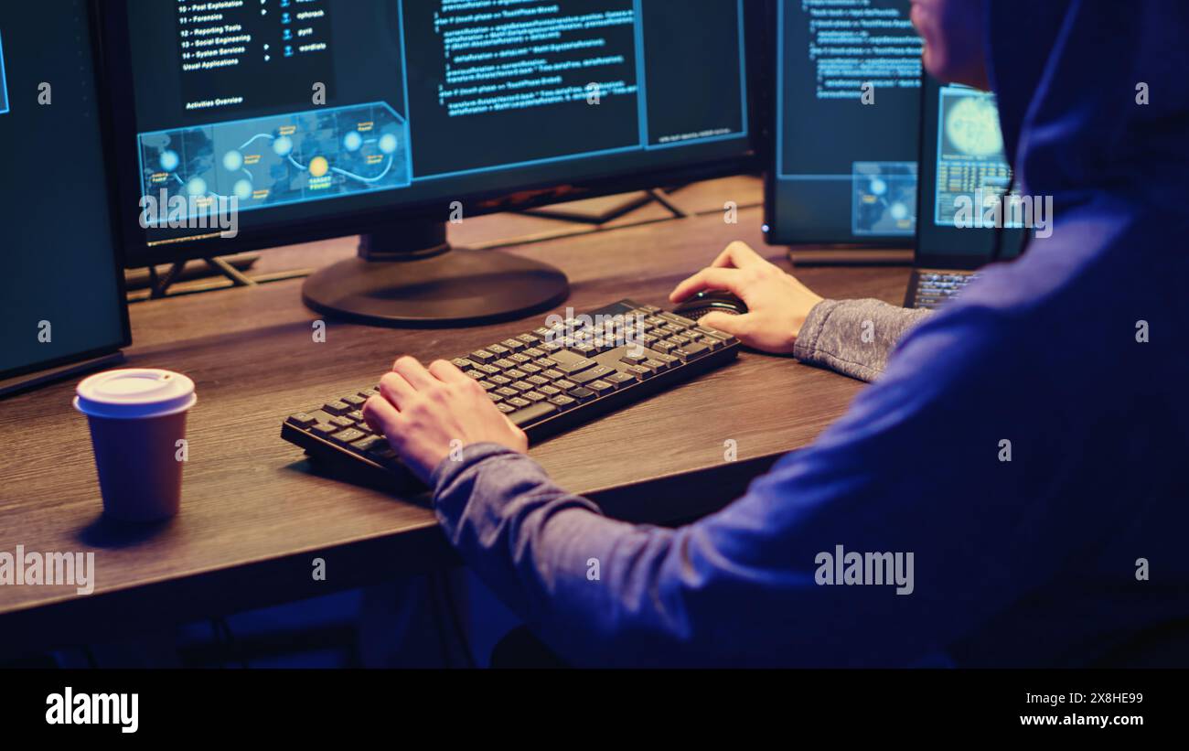 Young hacker drinking coffee while running spyware software designed to gather information from users computers without their knowledge. Script kiddie doing illegal stuff in graffiti painted hideout Stock Photo