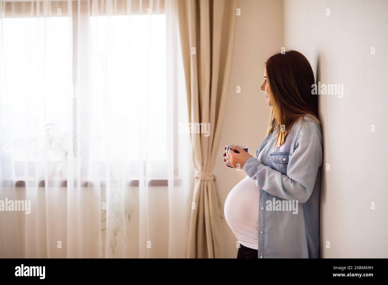 Pregnant woman leaning against wall in her home drinking herbal tea