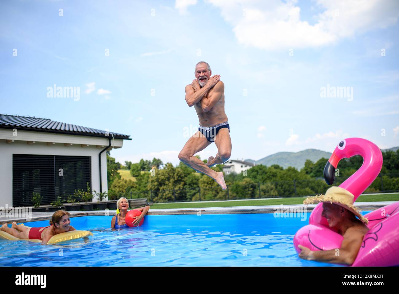 Group of cheerful seniors shaving fun in pool jumping, swiming and splashing. Elderly friends spending hot day by swimming pool. Stock Photo
