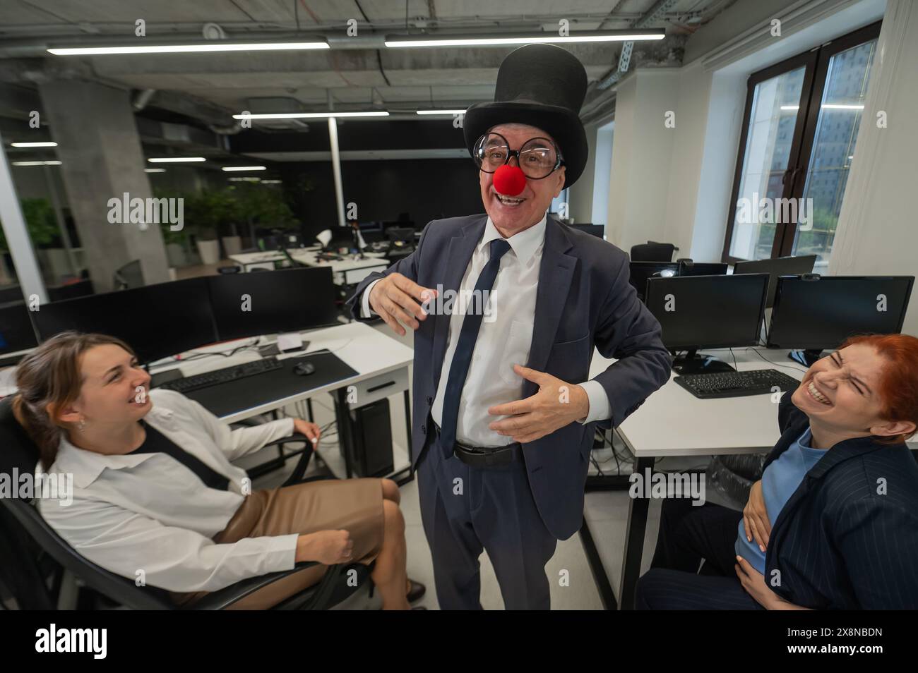 An elderly Caucasian man in a clown costume amuses two Caucasian women in the office.  Stock Photo