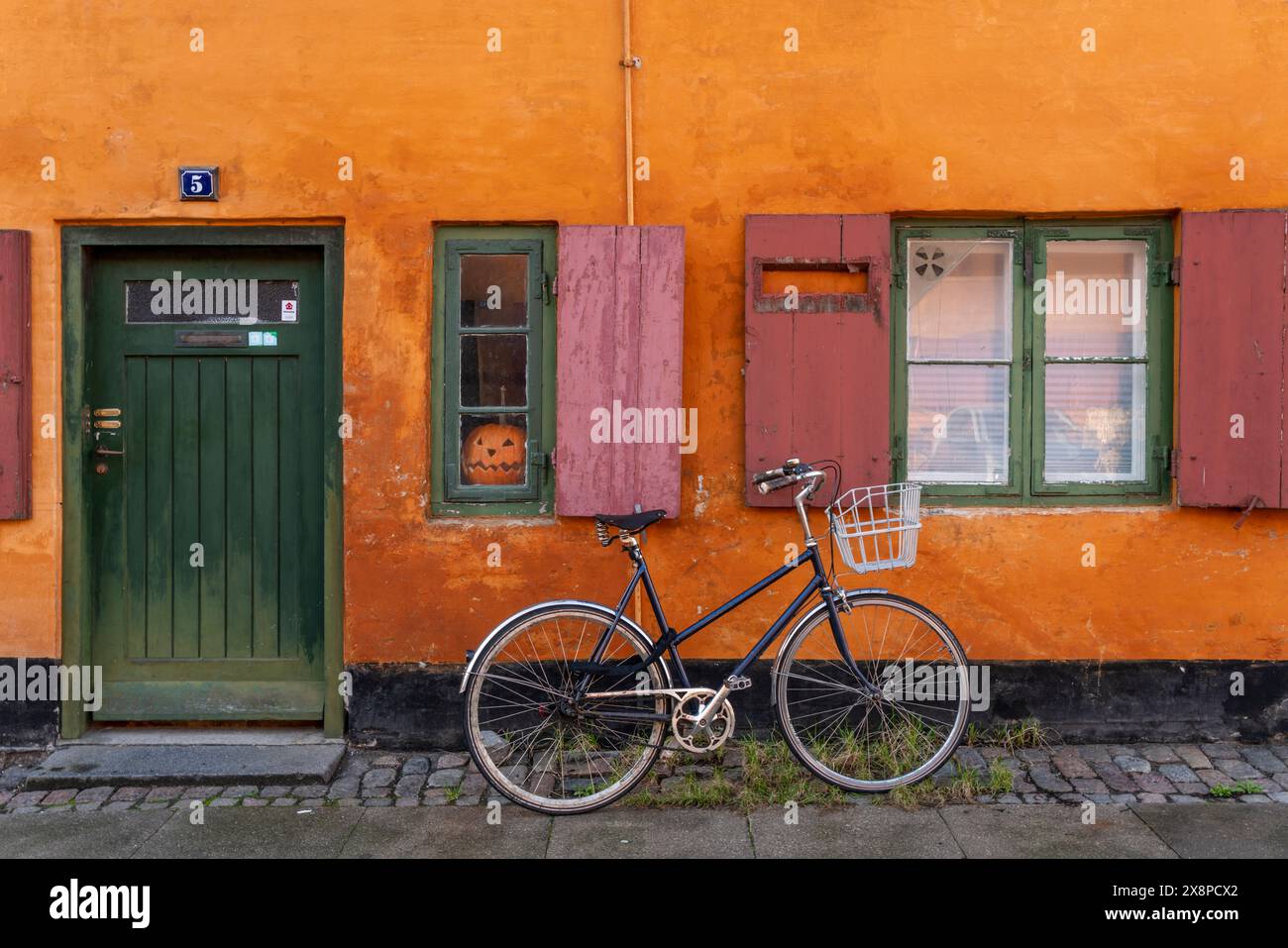 A picturesque European street comes to life in this image, where a vintage bicycle with a front basket leans against an orange wall Stock Photo