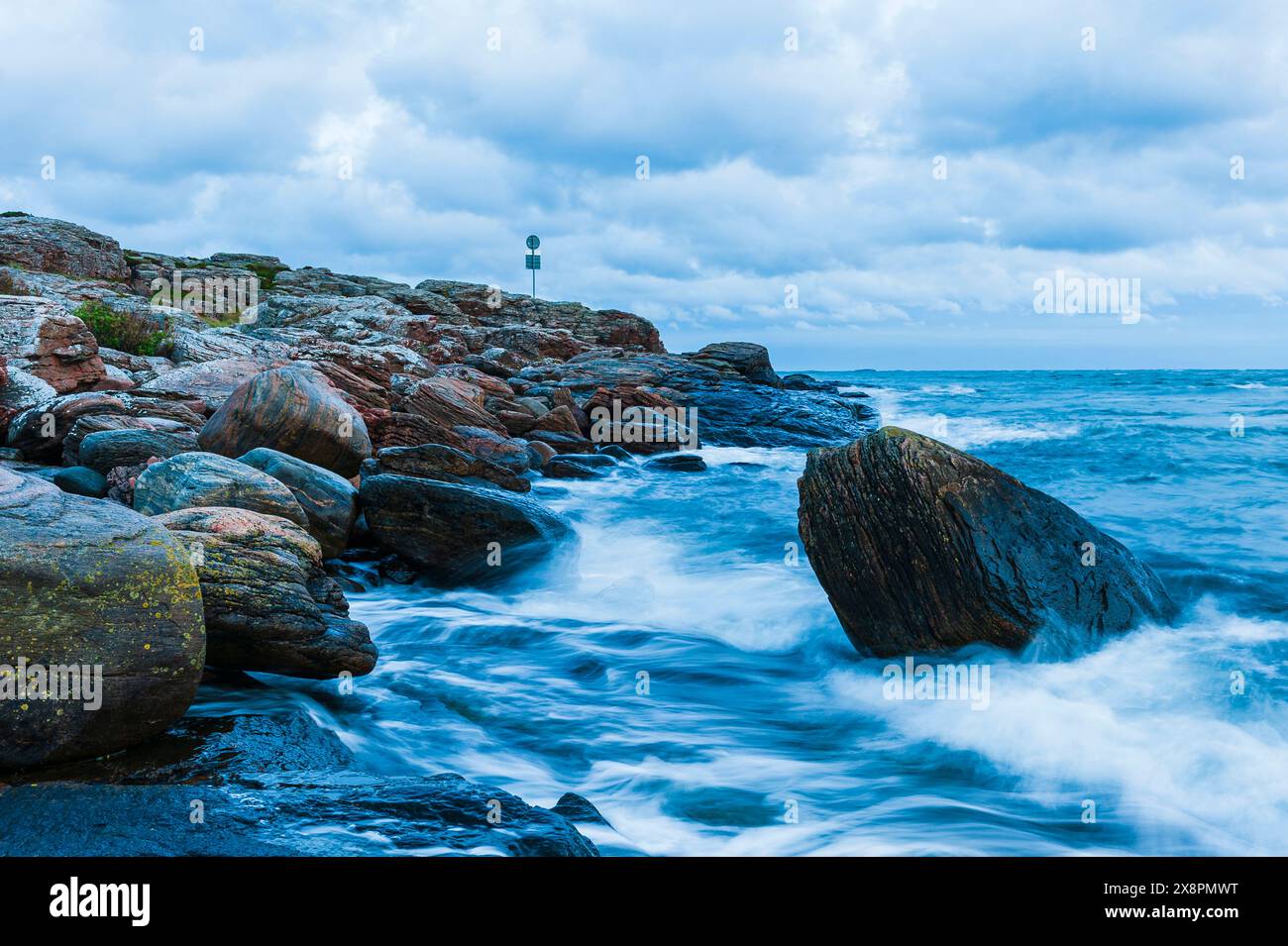 A rocky coastal shore in Sweden with a sign visible in the distance. The rugged rocks jut out towards the sea, and the lighthouse stands tall against Stock Photo