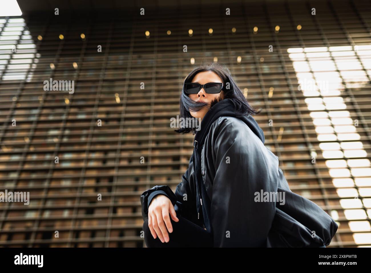 Young woman wearing sunglasses and a black hoodie is crouched down, looking confidently at the camera with a modern urban building in the backgr Stock Photo