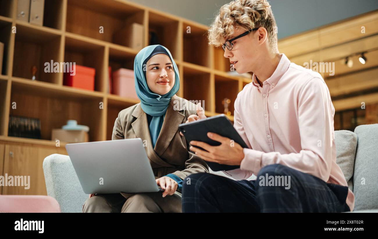 Two Creative Colleagues Discussing and Pointing at a Laptop in Modern Casual Office. Caucasian Male Sales Manager Discussing Project Plan on Tablet with Muslim Female Legal Assistant. Stock Photo