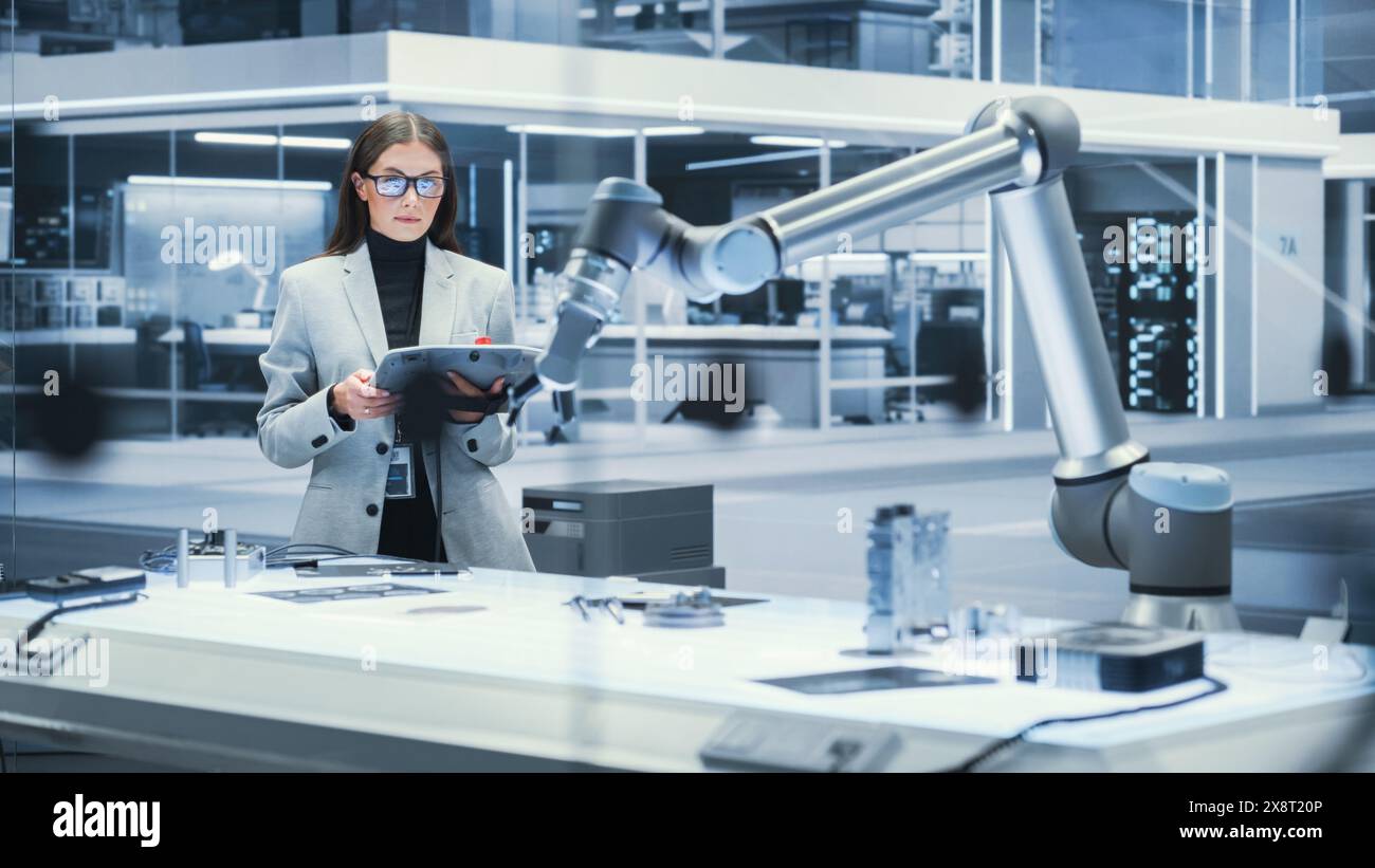Young Beautiful Female Engineer Using a Tablet Computer, Researching and Developing a Futuristic Robotic Arm Machine in a High Tech Industrial Laboratory with Modern Equipment. Cinematic Footage. Stock Photo