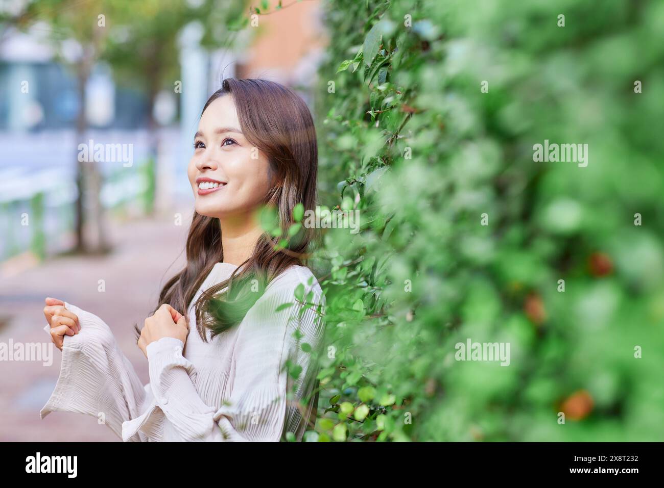 Japanese woman smiling while leaning against a wall with green plants ...