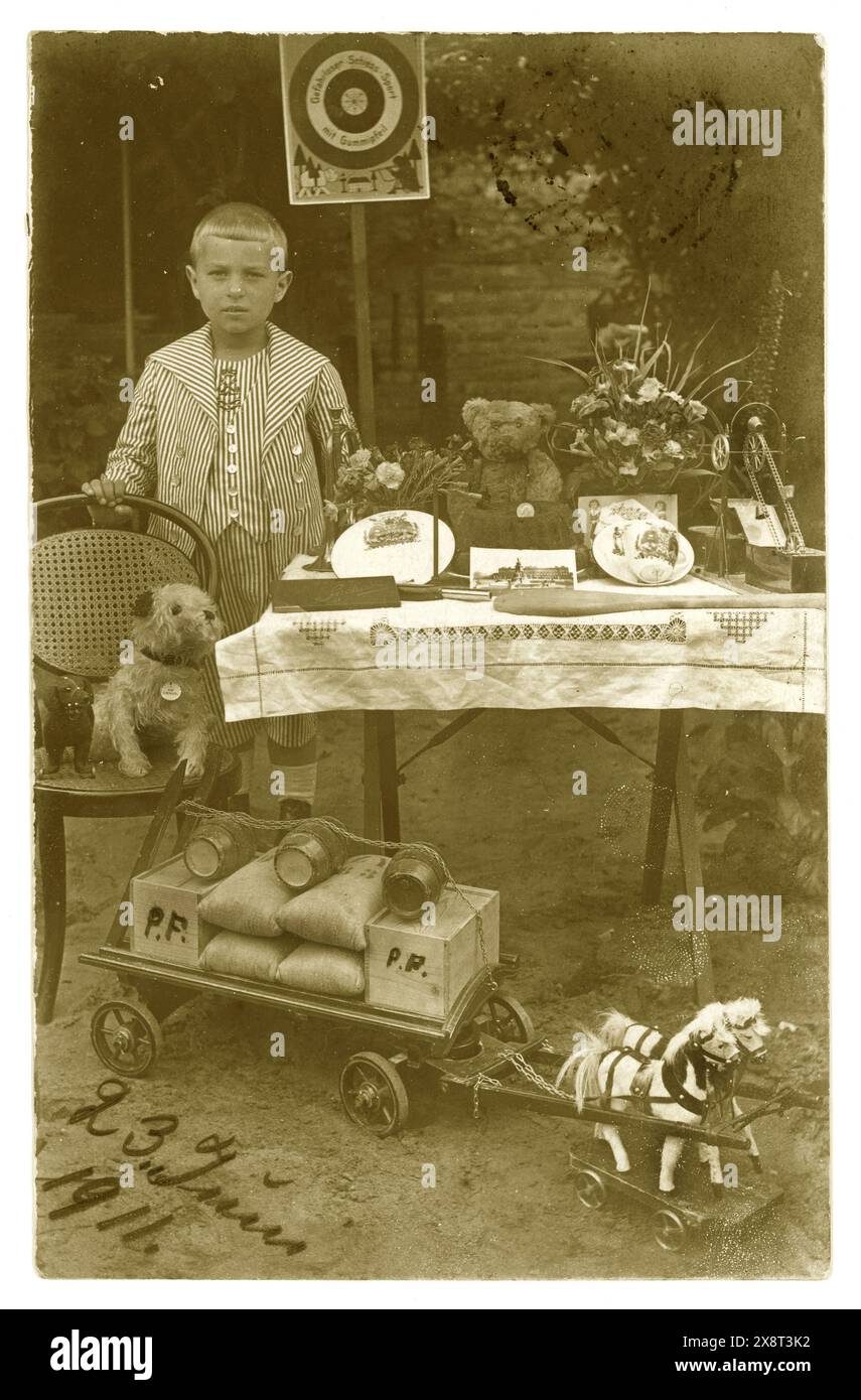 Original early 1900's postcard of young boy, wearing a smart sailor suit, displaying his toys outside in his garden, Birthday presents perhaps. Toys include teddy, soft toy dog, horse cart,  boy is Paul F maybe. Dated 23 June 1911 on front of postcard. Posted to London from Germany. Victorian boy boys /  Edwardian boy boys. Victorian child. Stock Photo