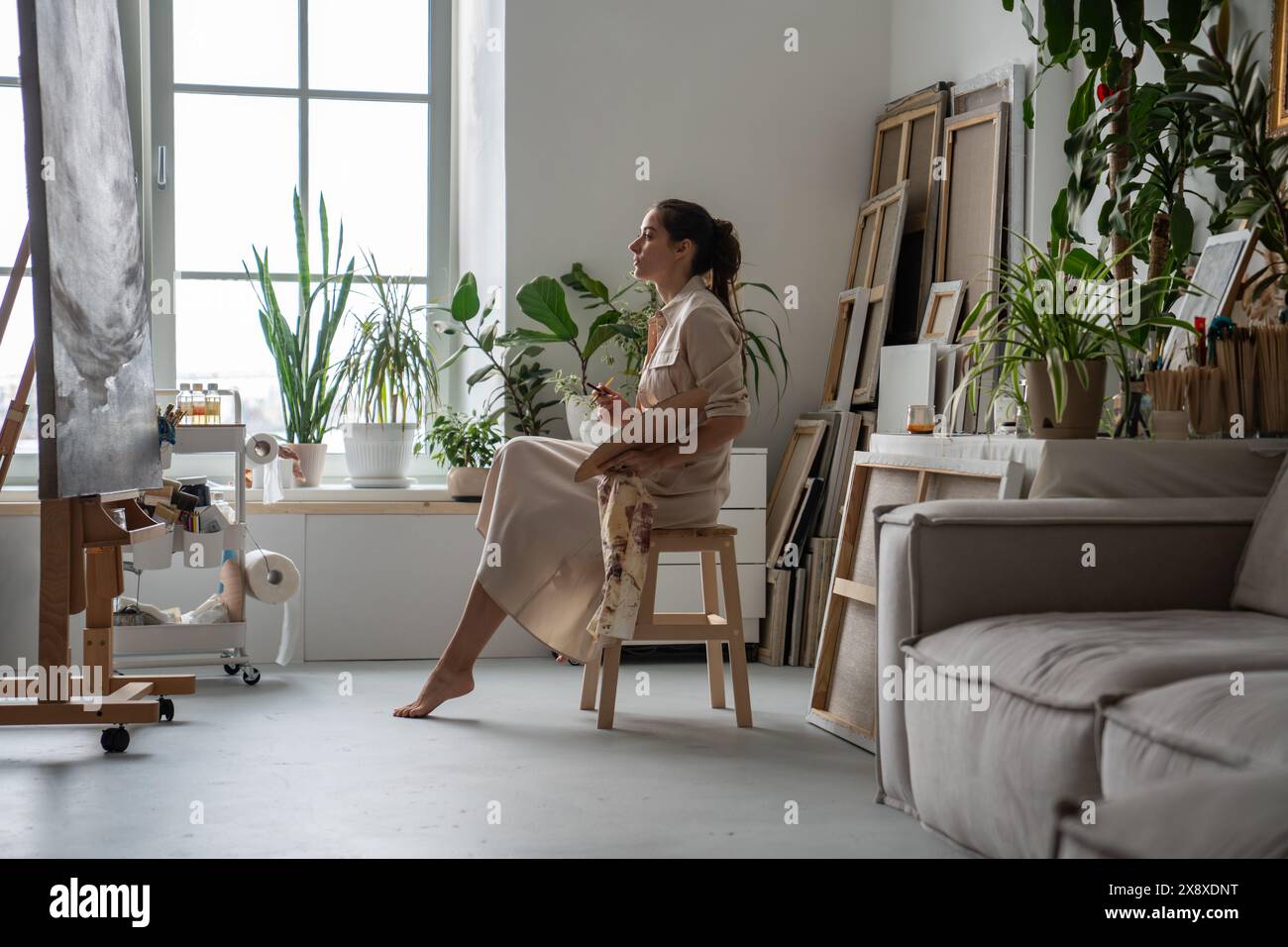 Thoughtful woman in art workshop studio sitting in front of large canvas with painting started. Stock Photo