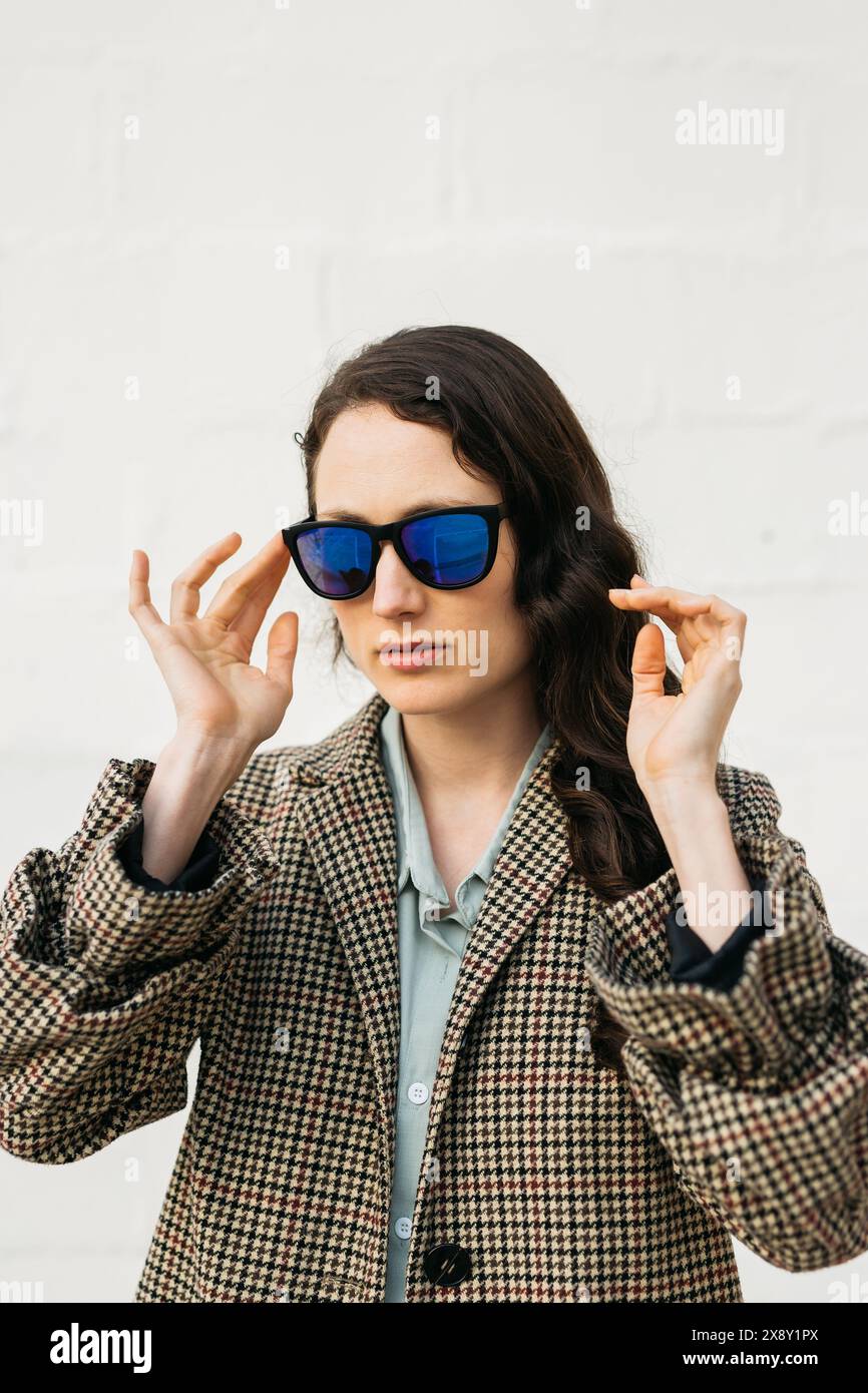 Portrait of a woman adjusting her sunglasses while wearing a houndstooth coat, standing against a plain white wall. Stock Photo