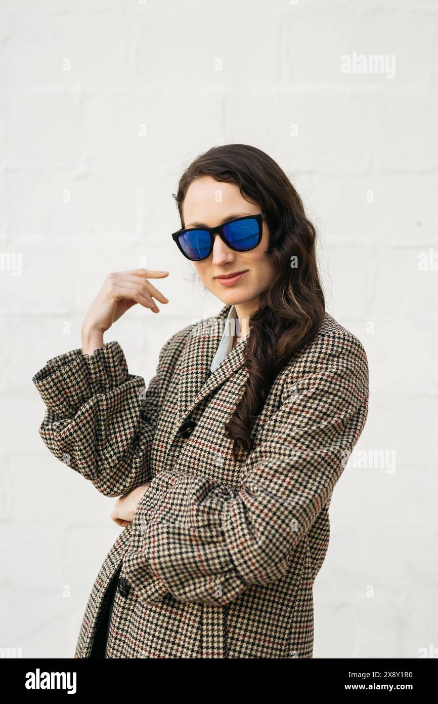 Portrait of a woman wearing sunglasses and a houndstooth coat, standing against a plain white wall. Stock Photo