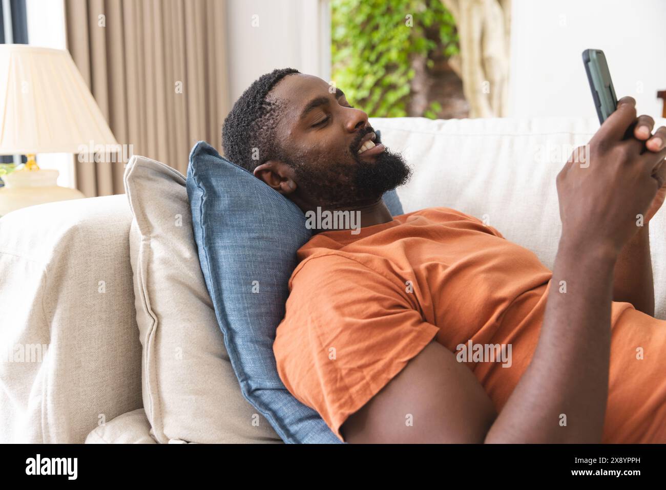 At home, young African American man lying on couch, laughing at smartphone Stock Photo