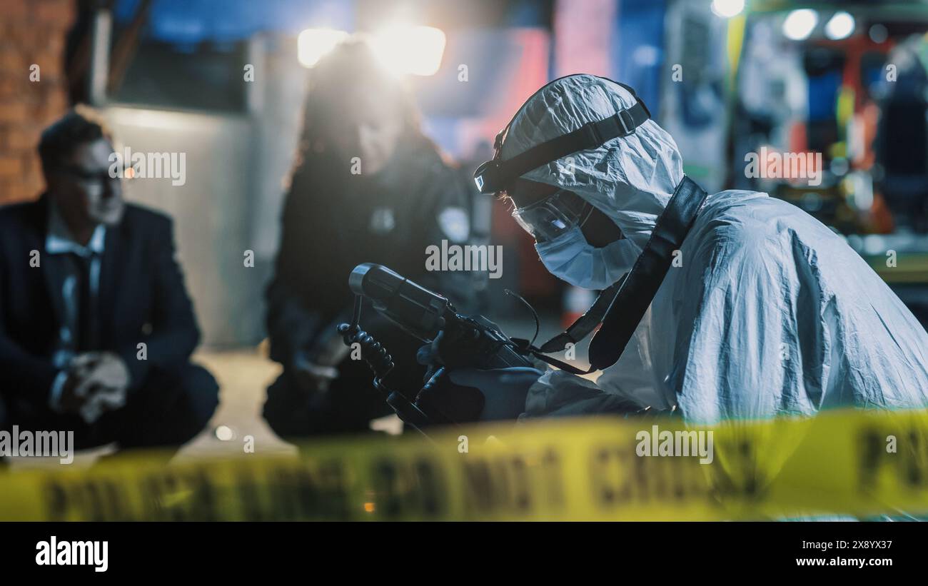 Forensics Specialist on Duty on Crime Scene at Night: Expert Using Advanced Camera to Take Photos of Evidence and Clues to Document Them for the Police Case File. CSI Team Working in the Background Stock Photo