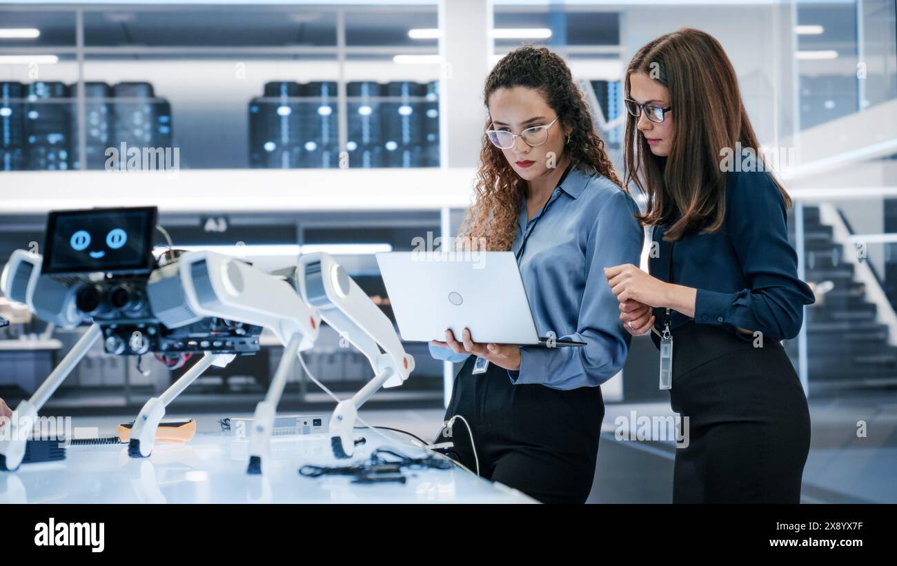 Two Industrial Robotics Engineers Gathered at a Table With Robotic Mobile Prototype. Two Females Discuss the Project, Program the Robot Dog with a Laptop Computer. Stock Photo
