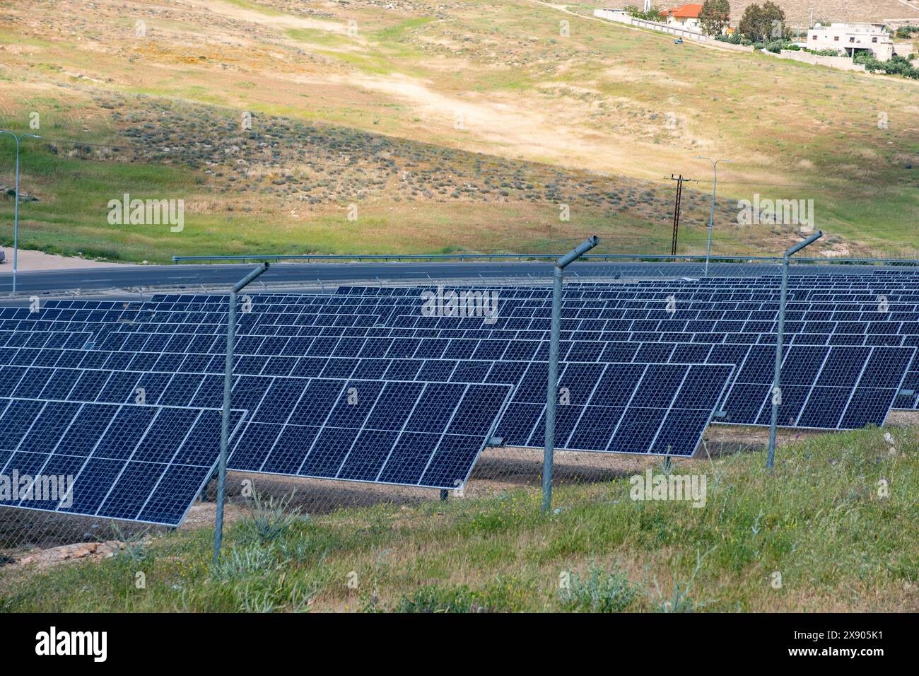 A vast array of solar panels is situated near a highway, harnessing solar energy in a rural landscape, with rolling hills and sparse buildings in the Stock Photo