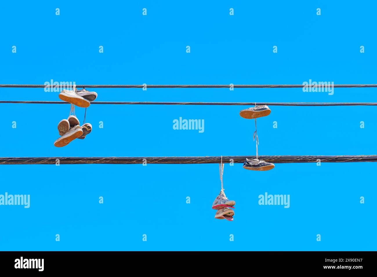 Several pairs of well-worn shoes are seen dangling from power lines, framed by a bright blue sky. Mysterious practice of shoe tossing concept. Stock Photo