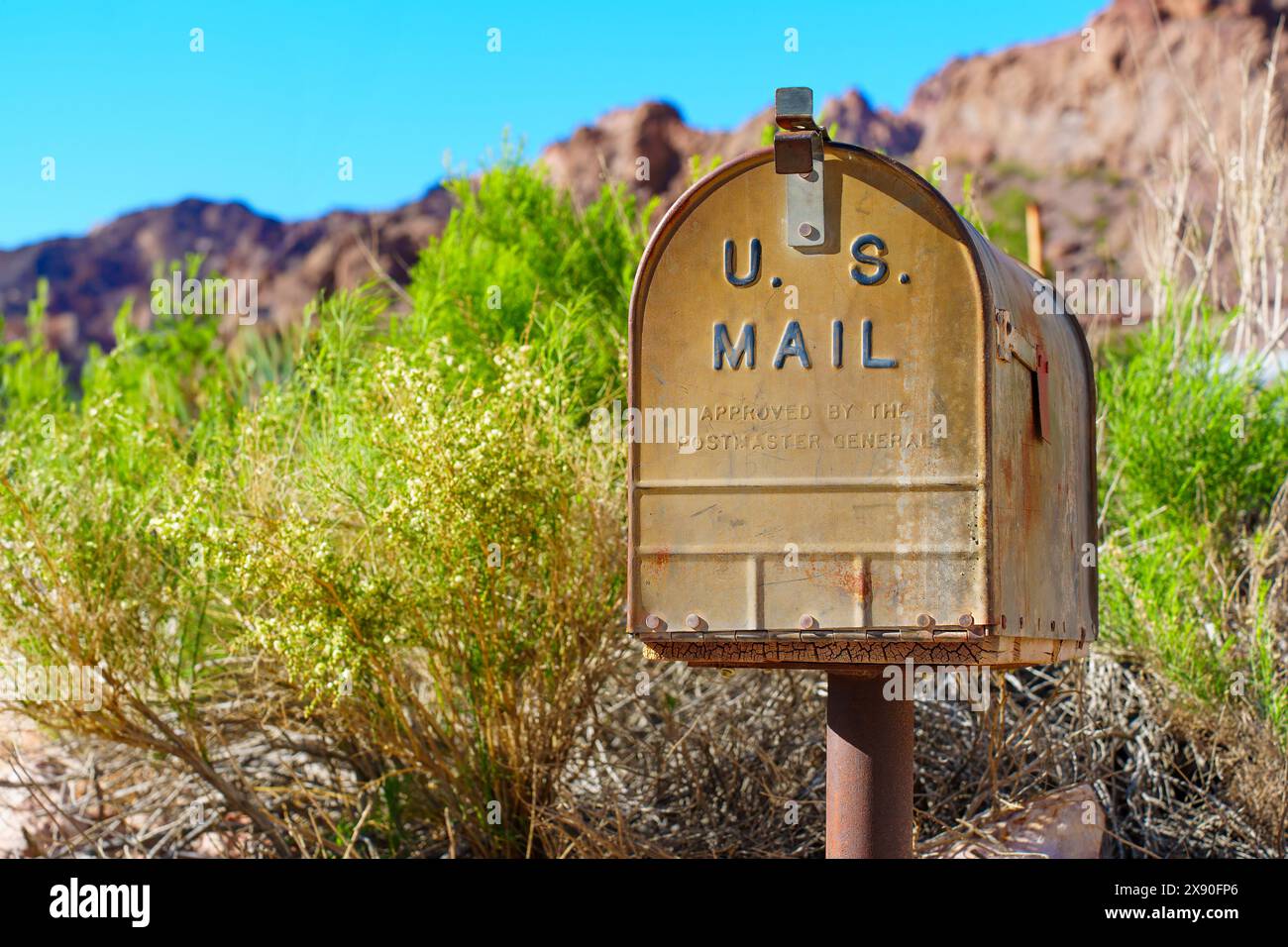 Nelson, Nevada - April 15, 2024: Rusted, metal U.S. Mail mailbox stands on a single post amidst green shrubbery with rocky terrain and a clear blue sk Stock Photo