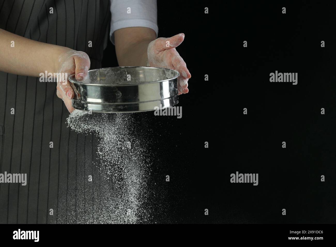 Woman sieving flour at table against black background, closeup. Space ...