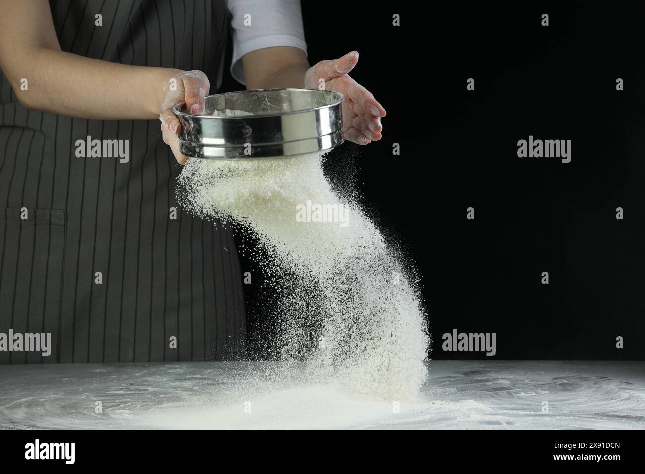 Woman sieving flour at table against black background, closeup. Space ...