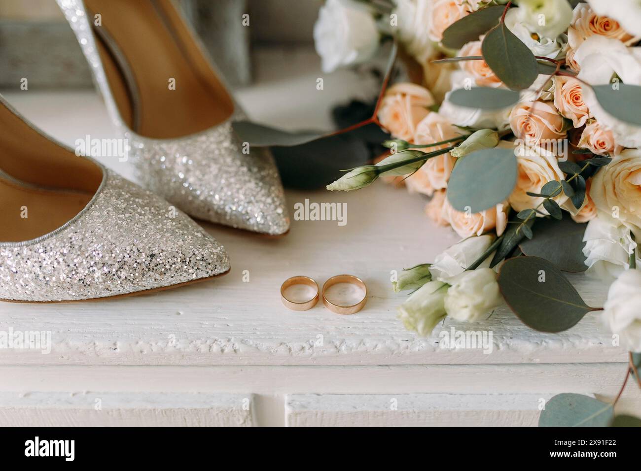 Elegant glitter shoes and wedding rings placed near a wedding bouquet of white flowers and peach roses with eucalyptus leaves on a wooden surface Stock Photo