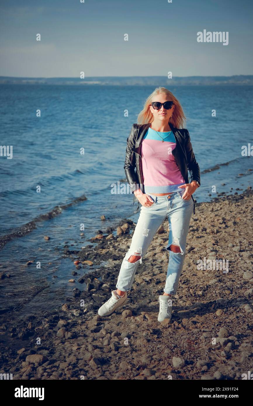 A woman wearing sunglasses, a leather jacket, and ripped jeans standing on a rocky beach, Belarus, Minsk Stock Photo