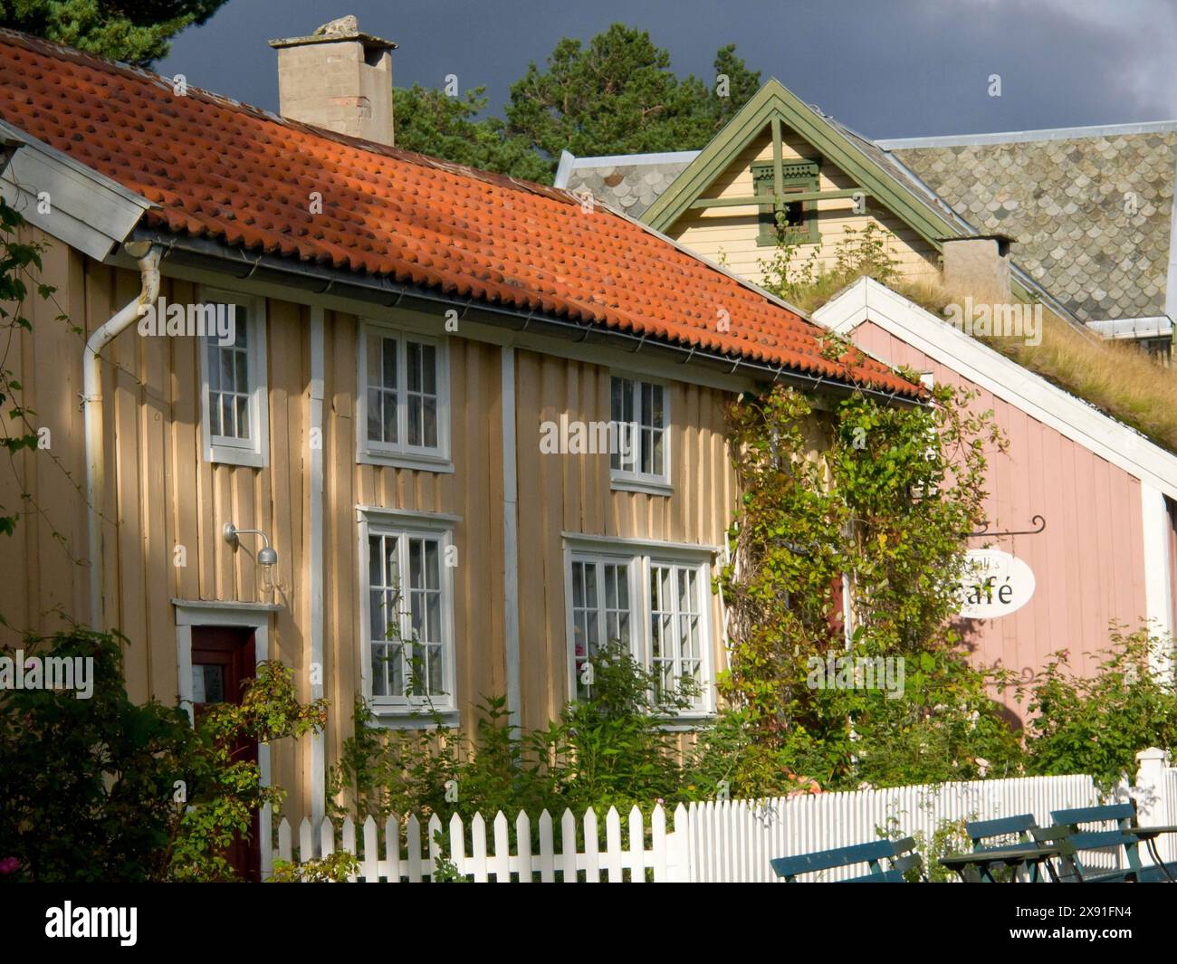 Yellow and pink house with a cafe sign and lush vegetation, colourful wooden houses behind white wooden fences, Molde, Norway Stock Photo