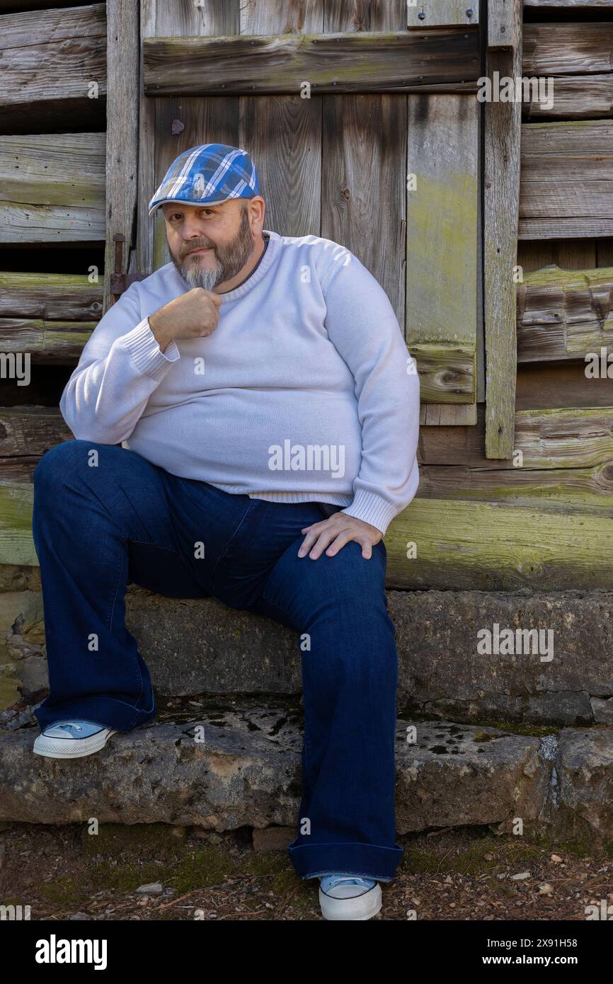 A man wearing a plaid blue cap, white sweater, jeans and tennis shoes with beard poses in front of an old rustic building. Stock Photo