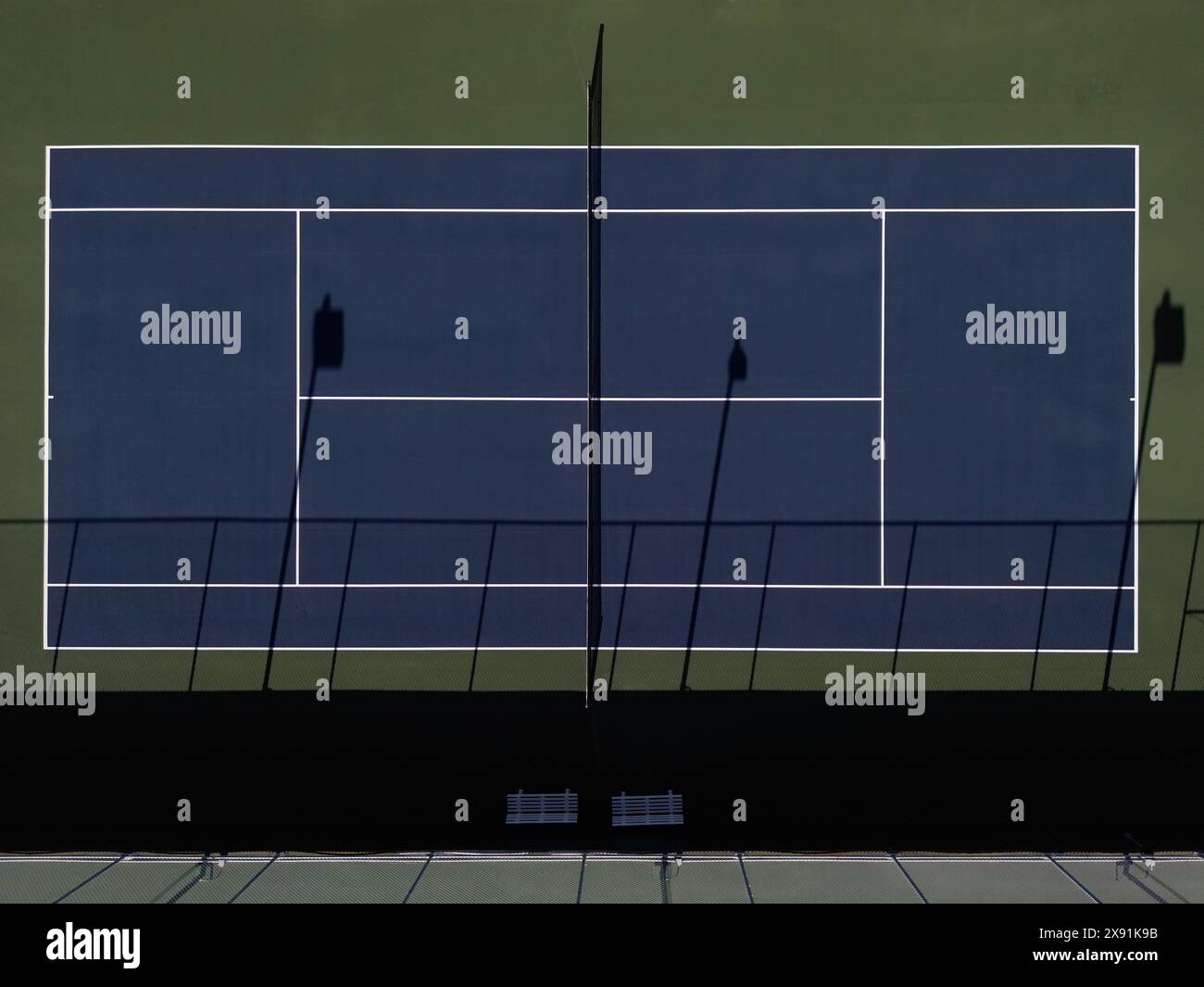 An early morning aerial view of a blue tennis court is shown, with shadows being cast on the playing surface. Stock Photo