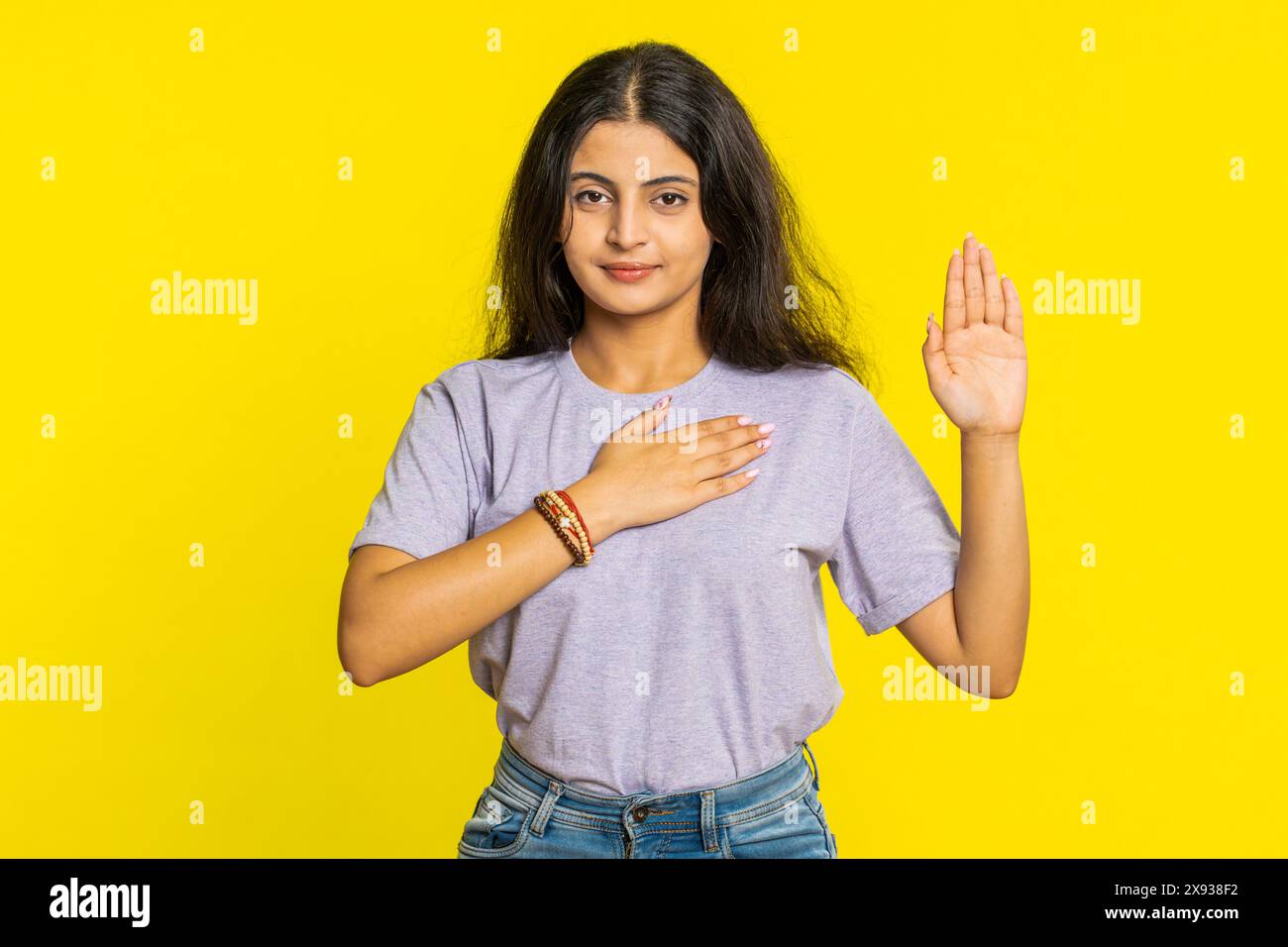 I swear to be honest. Sincere responsible Indian woman raising hand to take oath promising to be honest, to tell truth, keeping hand on chest justice. Arabian girl isolated on yellow studio background Stock Photo