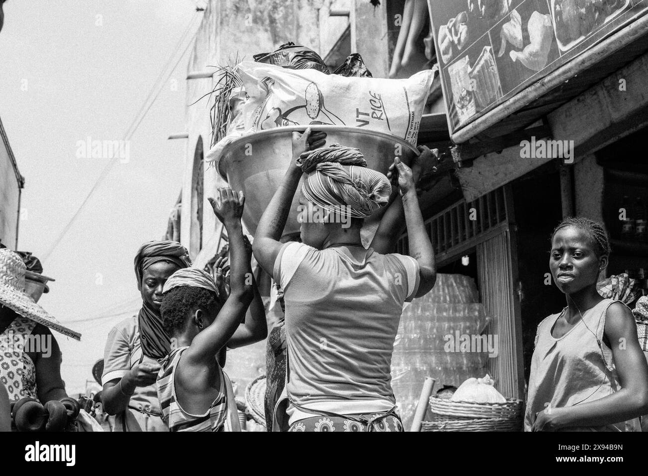 Women and children in a bustling market in Ghana, assisting with carrying heavy loads and engaging in market activities. Stock Photo