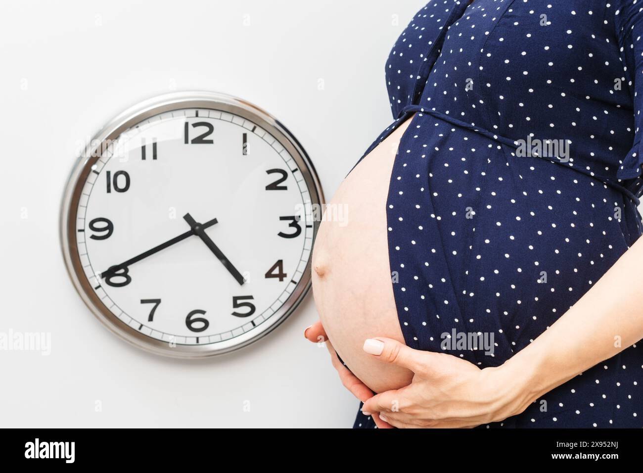 Pregnant woman holding her belly standing next to a wall clock during ...
