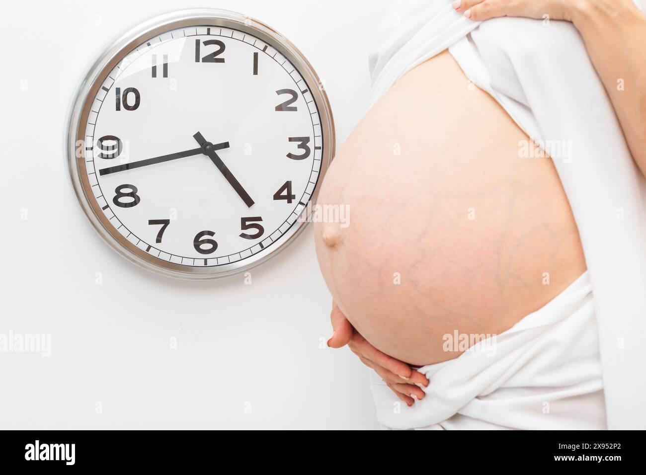 Pregnant woman holding her belly standing next to a wall clock during the final weeks of pregnancy Stock Photo