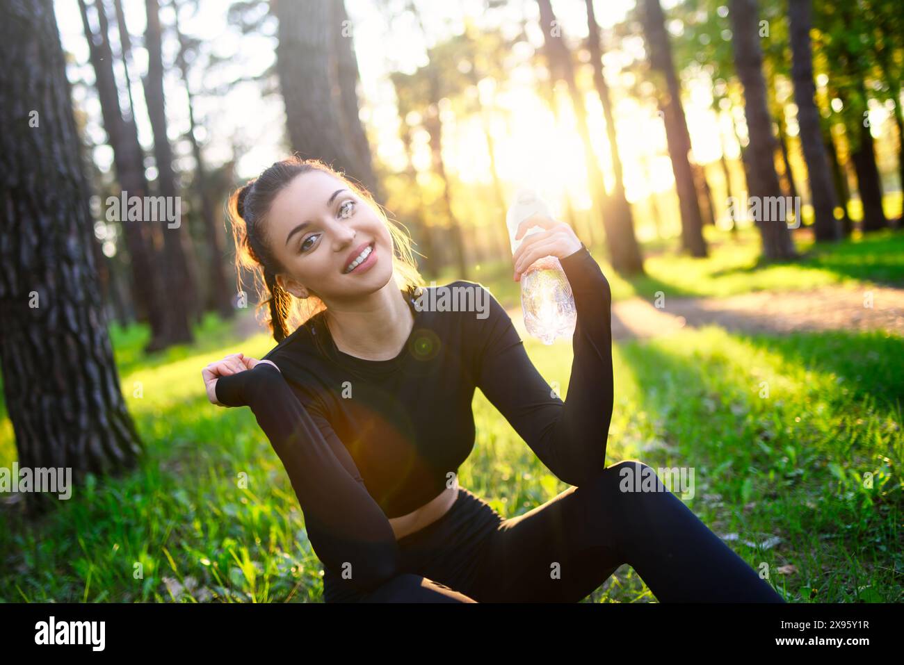 A smiling young woman in a sporty black suit holds a water bottle in the woods Stock Photo