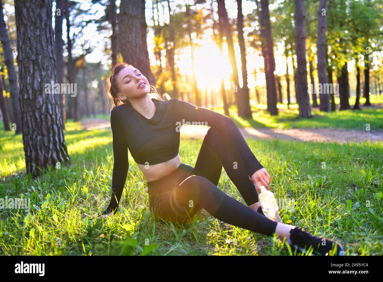 A smiling young woman in a sporty black suit holds a water bottle in the woods. Stock Photo