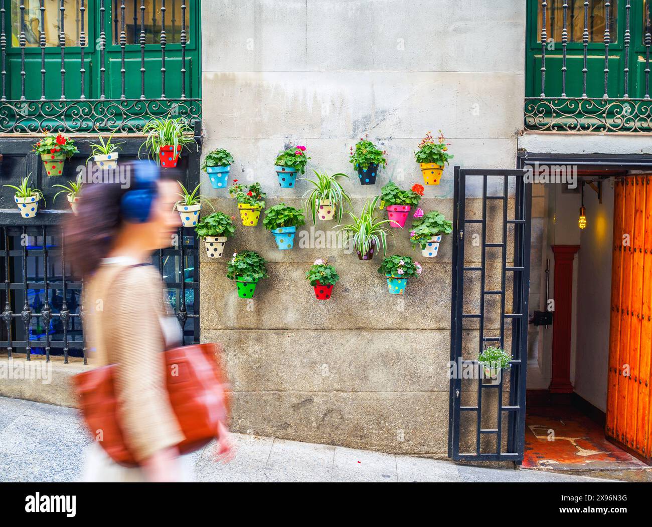 A girl with earphones walking past a wall adorned with colorful polka-dotted flower pots and plants. Stock Photo