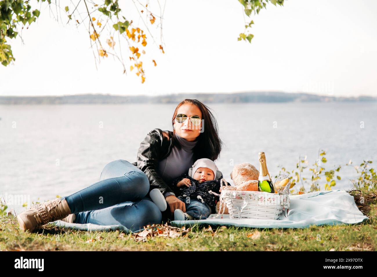 Woman with a baby lying on a blanket by a lake having a picnic, surrounded by autumn leaves Stock Photo