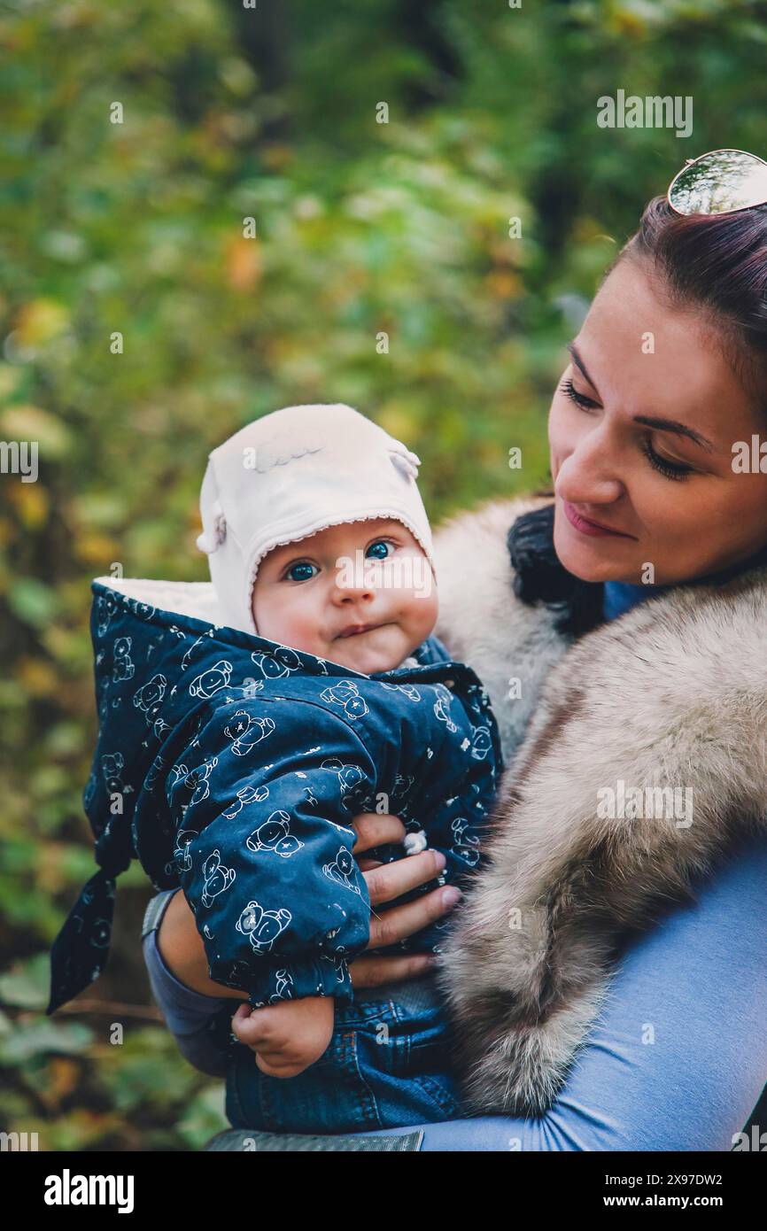 A woman holding a smiling baby in a blue jacket and white hat outdoors in a forested area Stock Photo