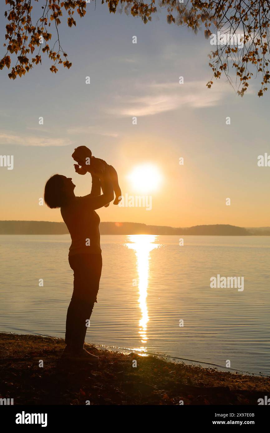 A silhouette of a parent lifting a child near a lake during sunset, reflecting on the calm water Stock Photo