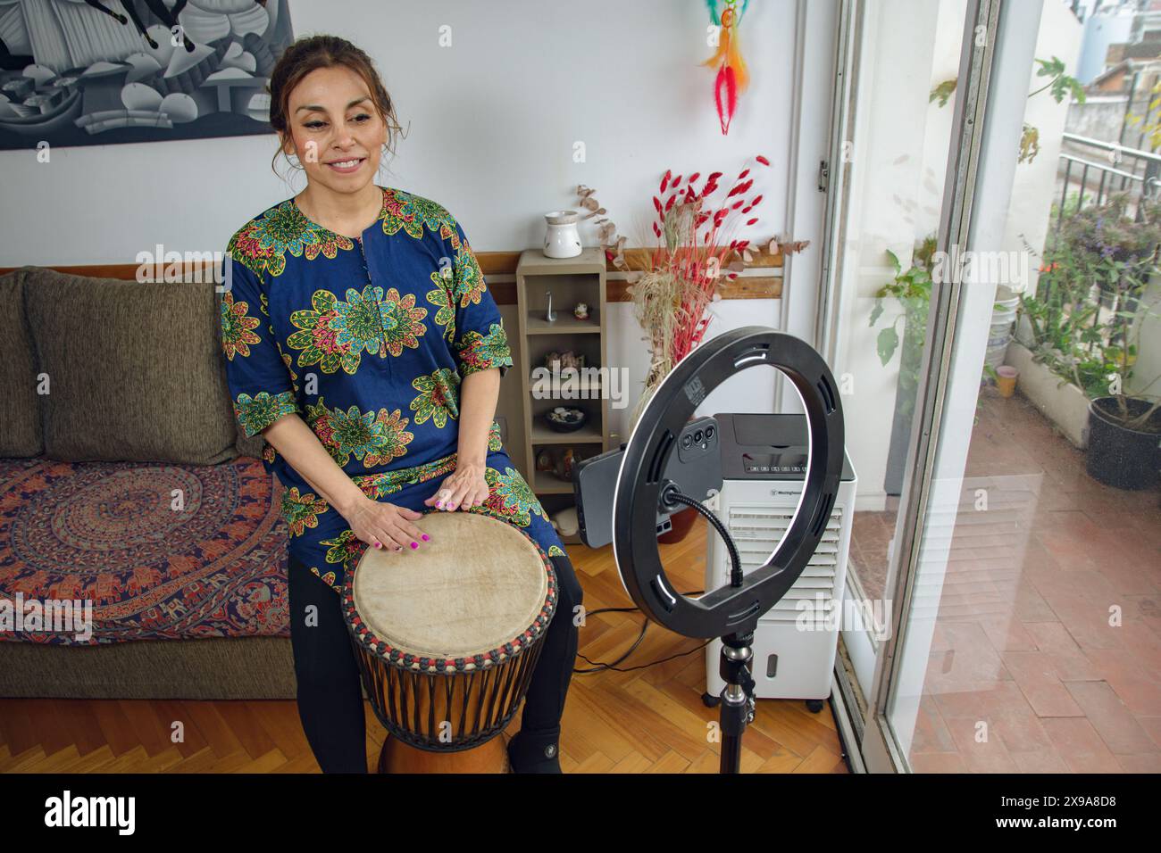 Adult Latin Woman enjoying the morning with her Djembe drum, making music and filming herself with a mobile phone. Is smiling happy and She is attenti Stock Photo