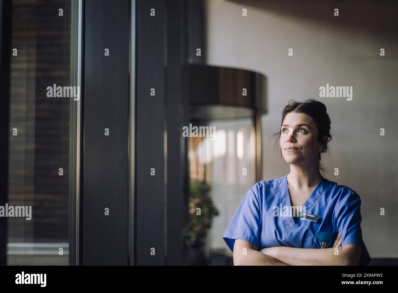 Thoughtful female physician in blue scrubs standing with arms crossed ...