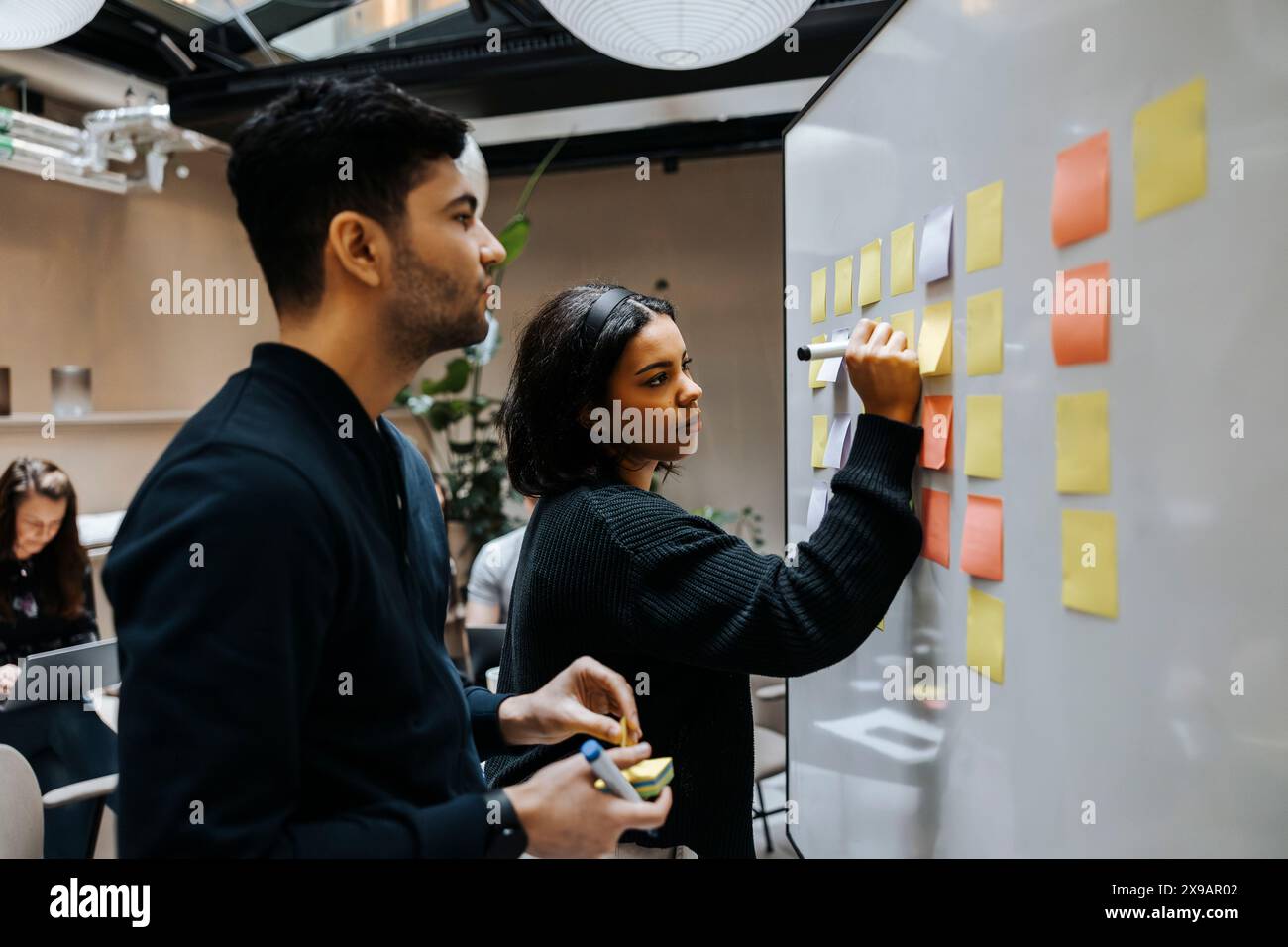 Female entrepreneur writing on adhesive note while standing next to male colleague near whiteboard at office Stock Photo