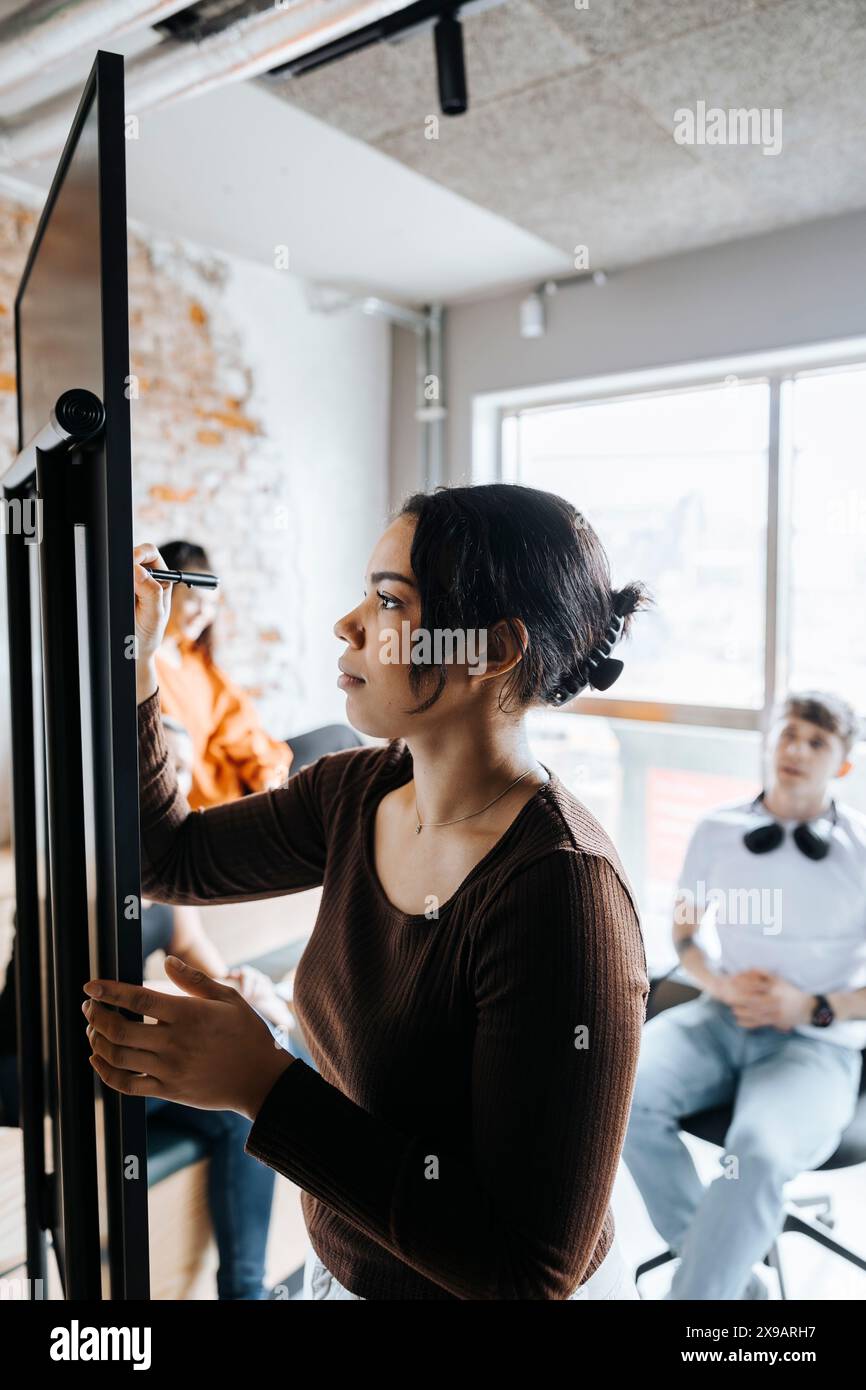 Young female entrepreneur writing on whiteboard in business meeting with colleagues at office Stock Photo