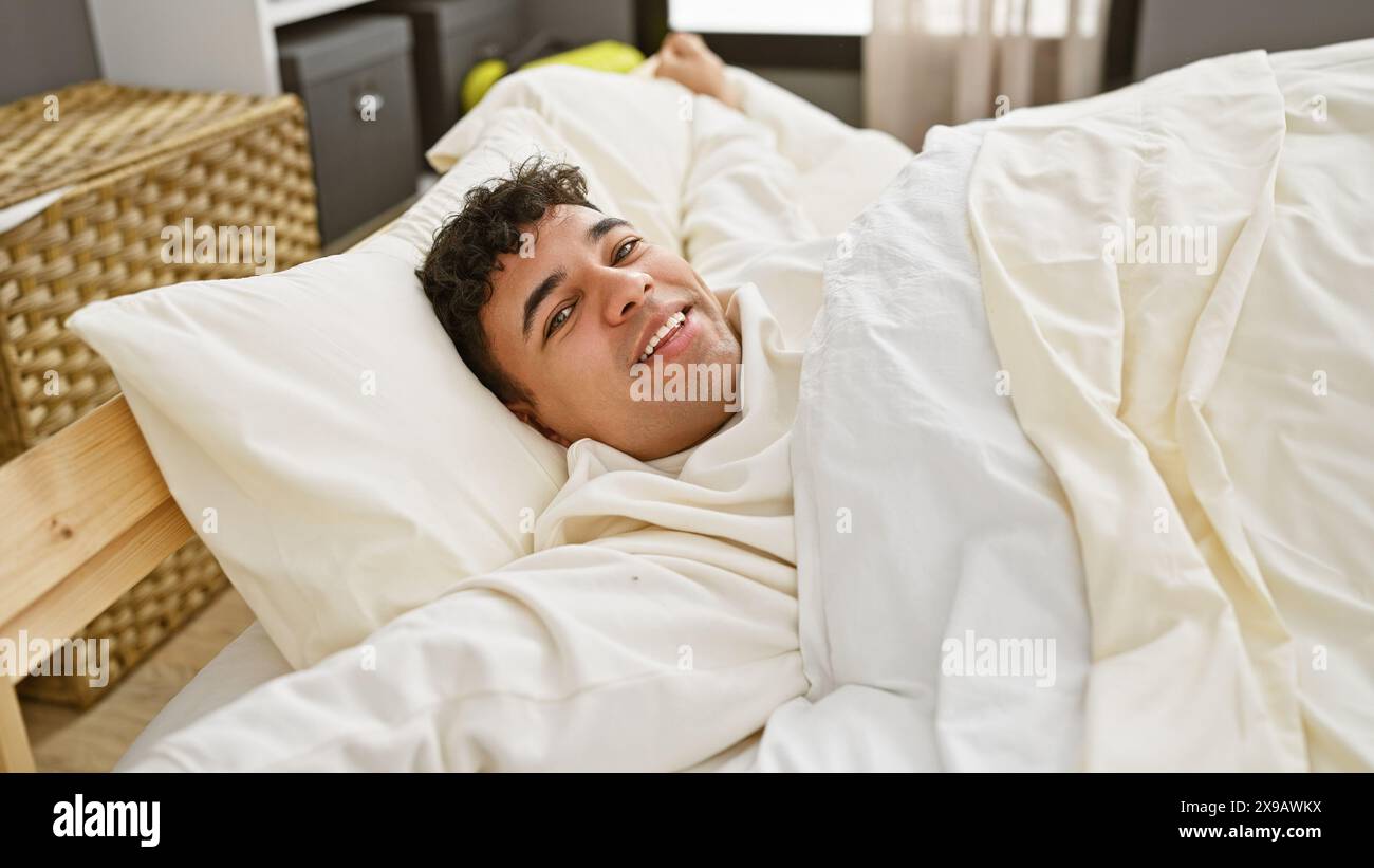 A cheerful young man with curly hair lying in bed, smiling in a bright bedroom setting. Stock Photo