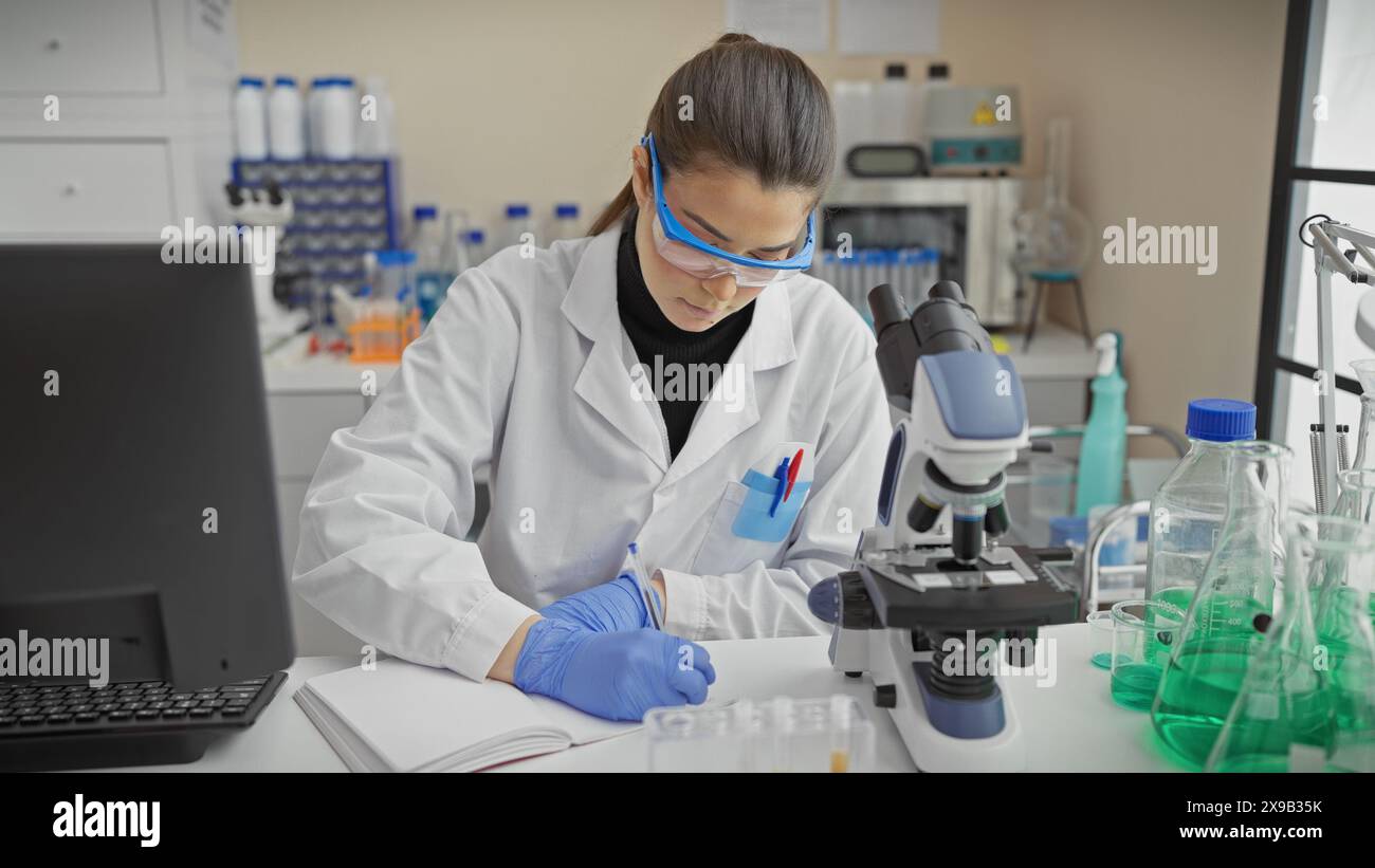 A focused hispanic woman wearing lab safety goggles works diligently in ...
