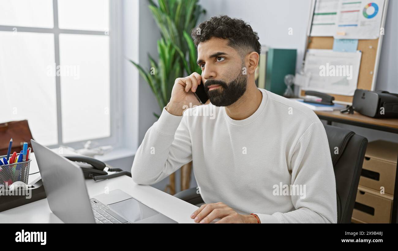 Handsome hispanic man with beard working on laptop in a modern office setting, looking pensive. Stock Photo