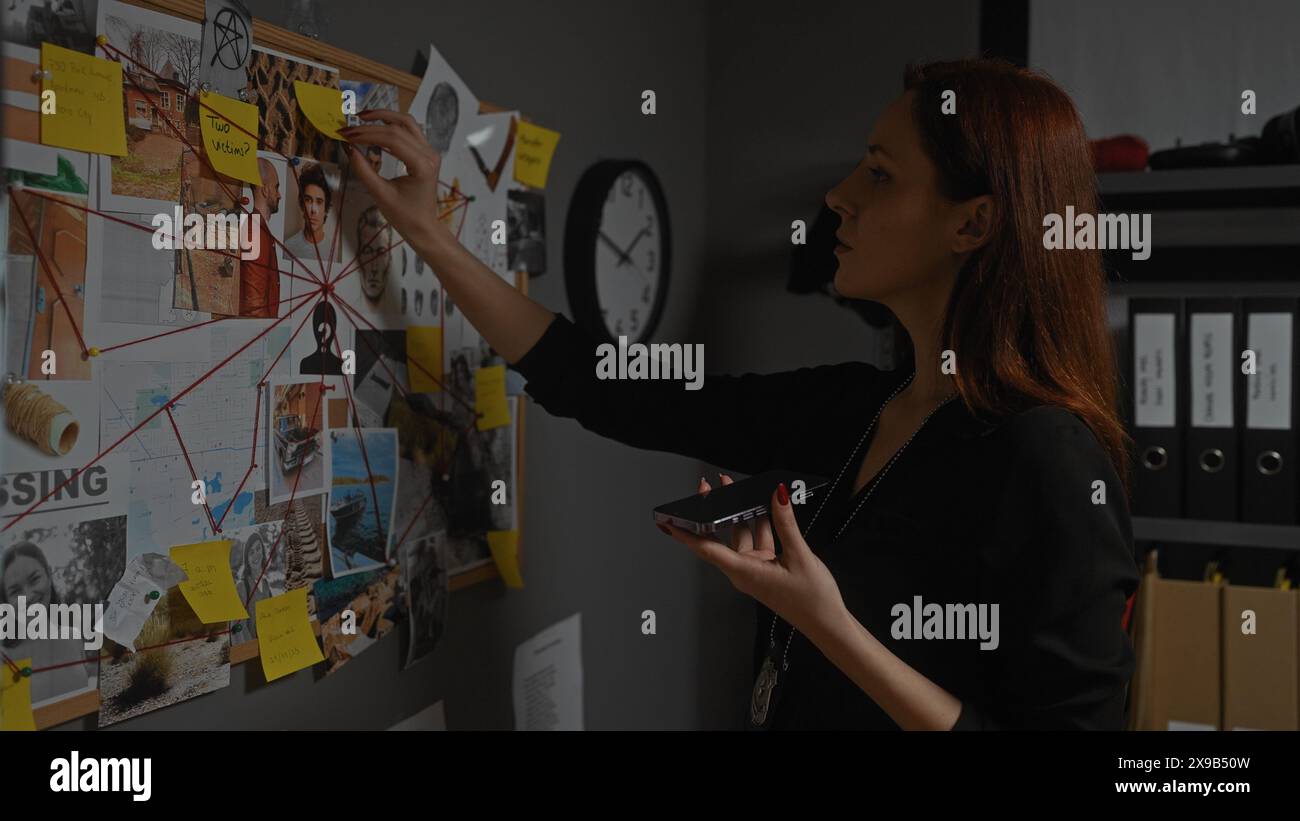 Redhead woman detective analyzing evidence board in office while holding phone Stock Photo